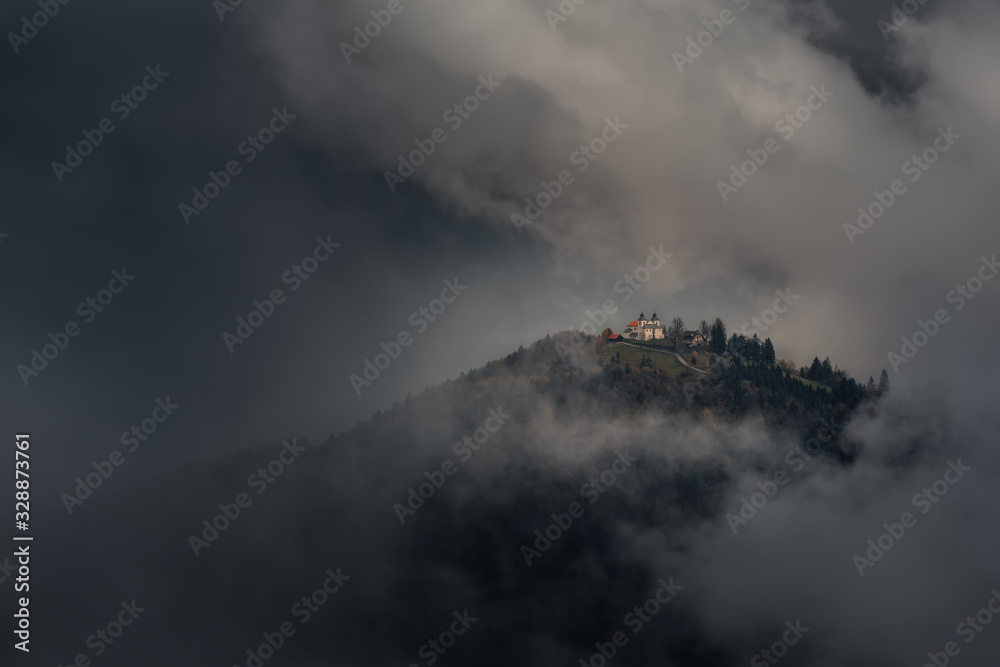Classic Autumn Slovenian Landscape In Dark Colors With A Small Church On Top Of A Mountain Among Thunderclouds. Shot Near The Famous Place Of Jamnik. Church With Red Tiled Roof In The Clouds