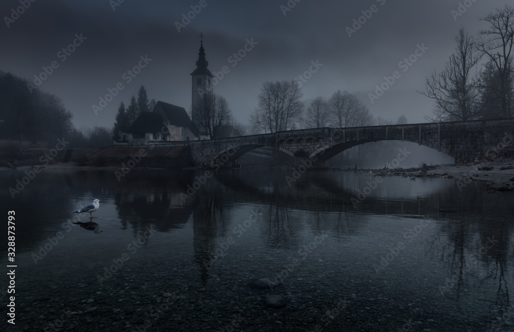 Autumn Dusk Foggy Landscape With Old Stone Bridge, Gothic Church of St. John the Baptist And Lonely Seagull. Lake Bohinj, National Park Triglav, Slovenia. Slovenian Landscape In Dark Colors