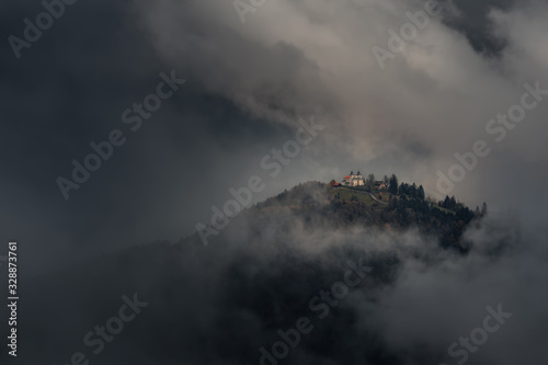Obraz na płótnie Classic Autumn Slovenian Landscape In Dark Colors With A Small Church On Top Of A Mountain Among Thunderclouds. Shot Near The Famous Place Of Jamnik. Church With Red Tiled Roof In The Clouds w salonie
