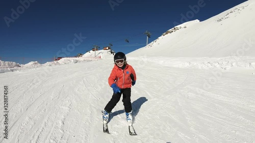Smiling child skiing and waving hand on snow mountain and blue sky background. Happy boy enjoying winter vacation on ski slopes in sunny day. Having fun with outdoors activity, sport