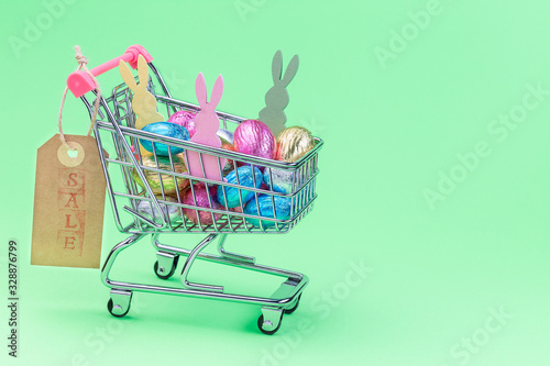 Green paper texture with shopping tray or basket full of colorful chocolate candy eggs and bunnies, paper tag with word sale, Easter background in pastel colors, horizontal, copy space