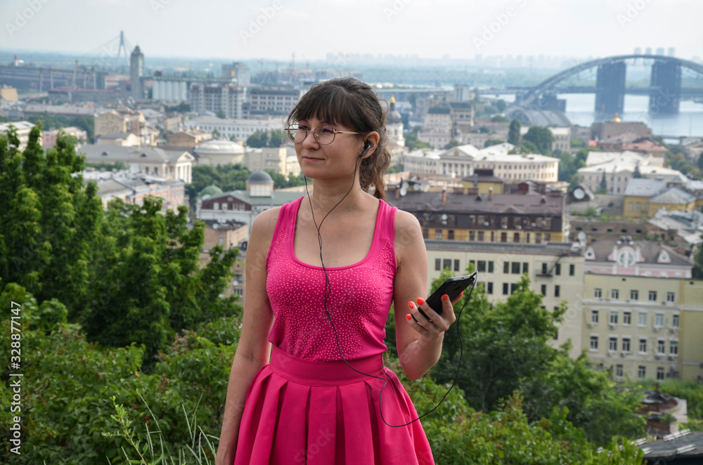 Portrait of beautiful attractive young girl in a pink dress and glasses with headphones and smartphone poses on a city background.