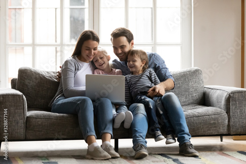 Happy young couple resting with children siblings on sofa, watching cartoons comedian movie on laptop. Overjoyed spouses having fun with joyful cute kids son daughter, using computer at home.