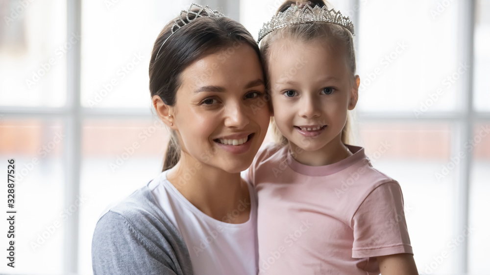 Close up head shot young mum embracing little cute blonde child daughter. Small pretty kid girl wearing crowns with affectionate babysitter nanny mother, looking at camera, happy family portrait. Close up head shot young mum embracing little cute blonde child daughter. Small pretty kid girl wearing crowns with affectionate babysitter nanny mother, looking at camera, happy family portrait.