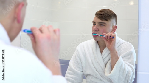 A young man is brushing his teeth. A man in a white coat started brushing them with a toothbrush. View through the mirror