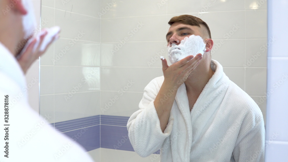 A young man applies shaving foam in front of a mirror. A man in a white coat with foam on his face. View through the mirror.
