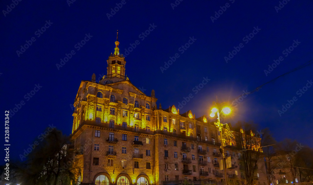 A tall building captured at night. The whole building is lighted with warm yellow light, making it look golden. It contrast with dark navy blue sky. A tall tree growing in front of it.