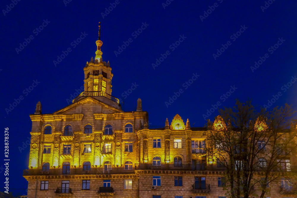 Obraz premium A tall building captured at night. The whole building is lighted with warm yellow light, making it look golden. It contrast with dark navy blue sky. A tall tree growing in front of it.