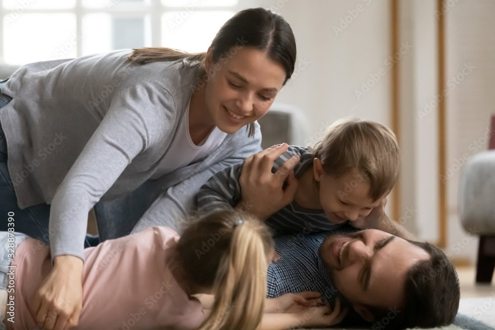 Playful family of four lying on floor, having fun together in living room. Happy married couple playing with joyful laughing little children siblings, enjoying free weekend active time indoors