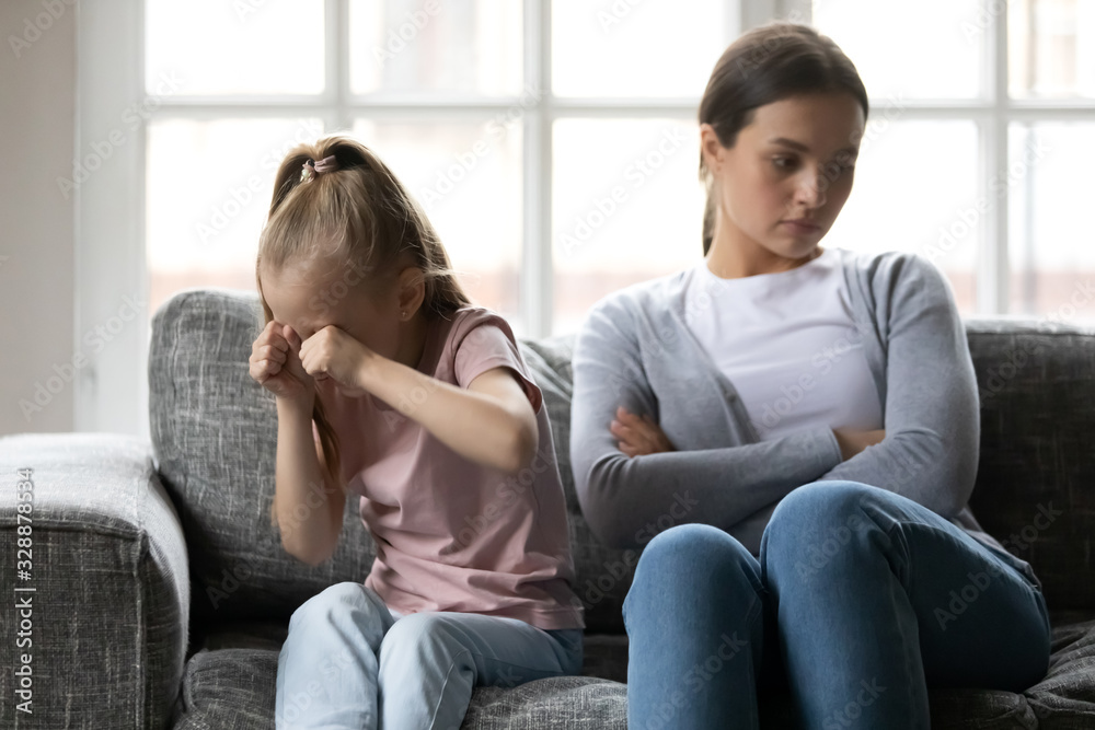 Stressed little school aged girl crying, abused by frustrated young nanny. Unhappy young mother ignoring small desperate child daughter, sitting together on comfortable couch in living room.
