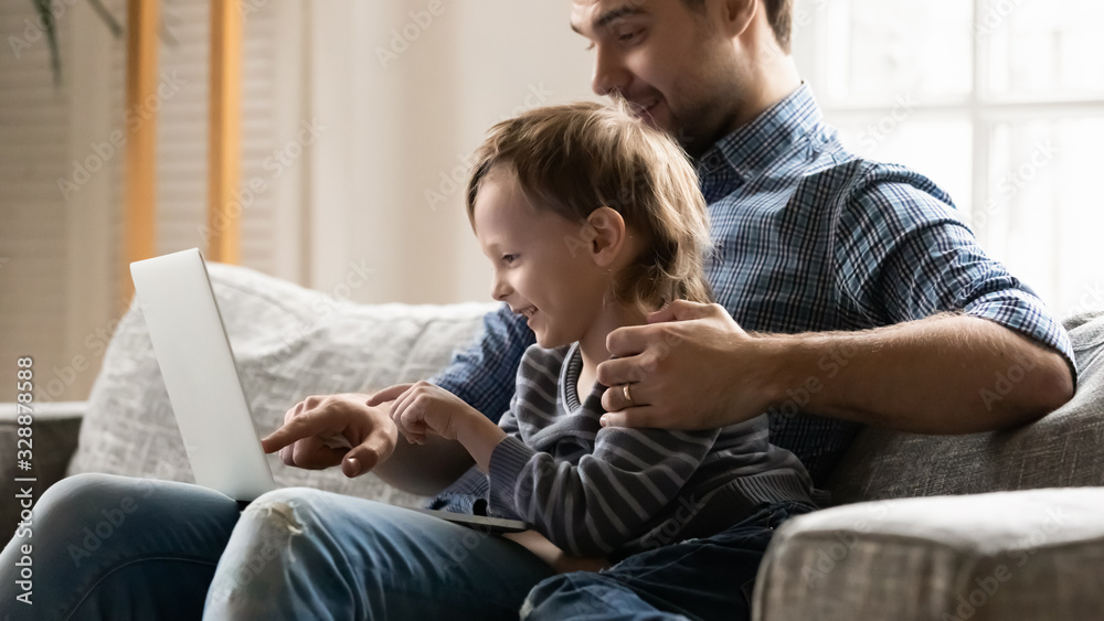 Side view smiling young father showing educational app to little kid son on computer. Happy small child boy enjoying playing online game or shopping together on laptop with joyful dad at home.