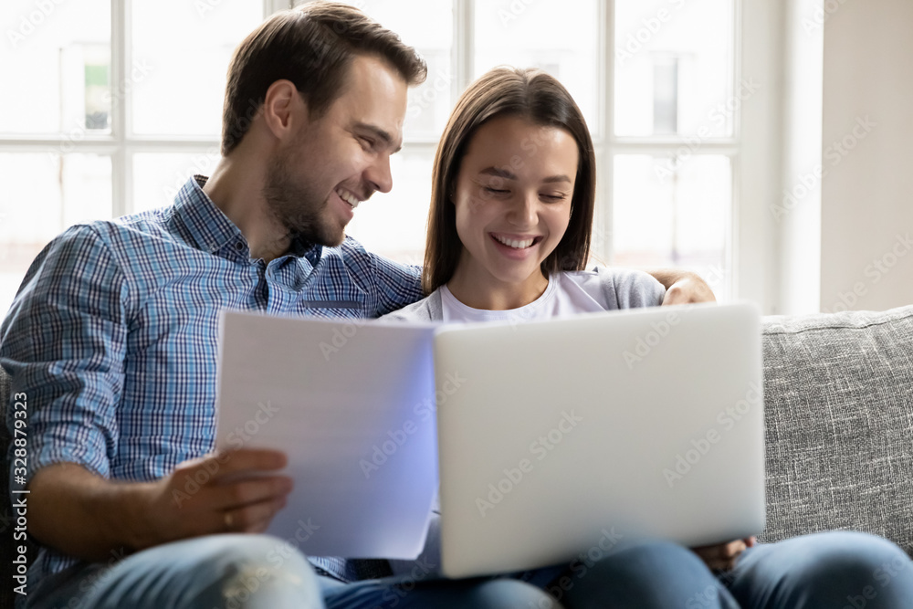 Smiling young man holding papers, embracing happy wife, using computer accounting applications at home. Joyful family couple filling banking information for loan mortgage payment in living room. Smiling young man holding papers, embracing happy wife, using computer accounting applications at home. Joyful family couple filling banking information for loan mortgage payment in living room.