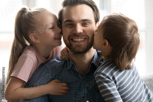 Head shot close up portrait cheerful little kids siblings kissing cheeks of happy young father. Smiling dad enjoying sweet tender moment with cute small children son daughter, looking at camera.