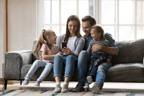 Full length joyful family of four sitting on couch, watching funny video together at home. Smiling young woman taking selfie photo with affectionate husband and cute kids son daughter on smartphone.