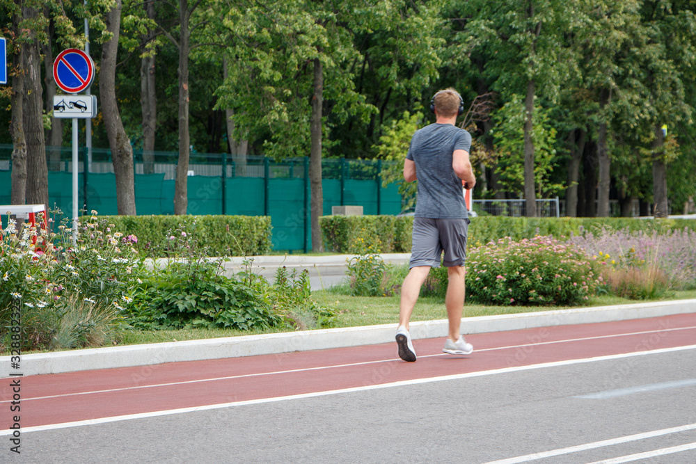 A young man in a gray T-shirt and shorts is running with headphones on a track in a city park.