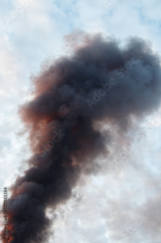 Black column of smoke in the cloudy sky. Orange reflexes of sunset on a pillar of smoke.