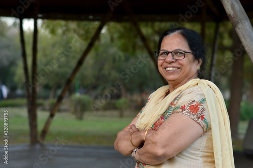 Smart and confident senior north Indian woman standing, posing for the camera with hands crossed / folded in a park wearing an off white salwar kameez punjabi suit in summers in New Delhi, India photo