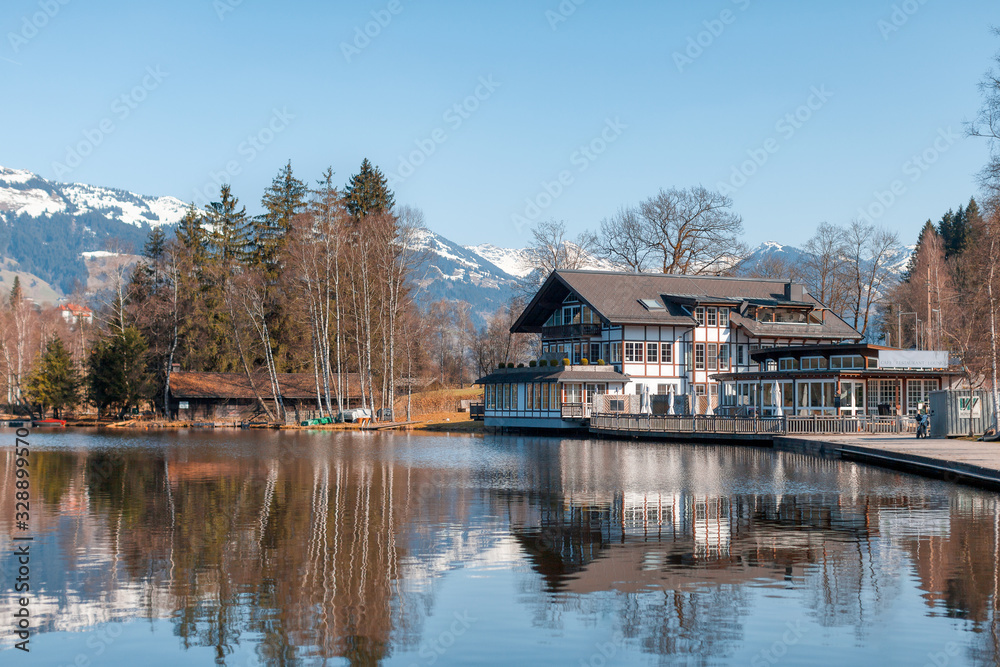 Cozy wooden house/chalet on the lake coast. Vacation in Austrian Alps. Snowy mountains in background. Tourism concept. Puddle reflection.
