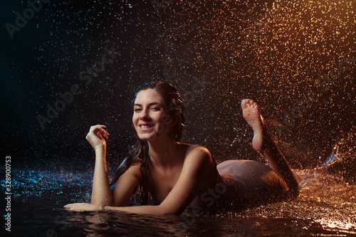 Beautiful sexy girl with drops of water on her face in splashes illuminated by an orange light against a dark background in the studio.