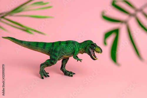 Fototapeta Naklejka Na Ścianę i Meble -  green dinosaur toy with open mouth between blurred palm leaves on a pastel pink  background