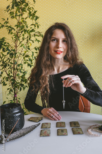 Tyumen, Russia, 12 February 2020 - beautiful young woman in a black velvet dress holds a crystal on a silver chain above the runes during a spiritual seance photo