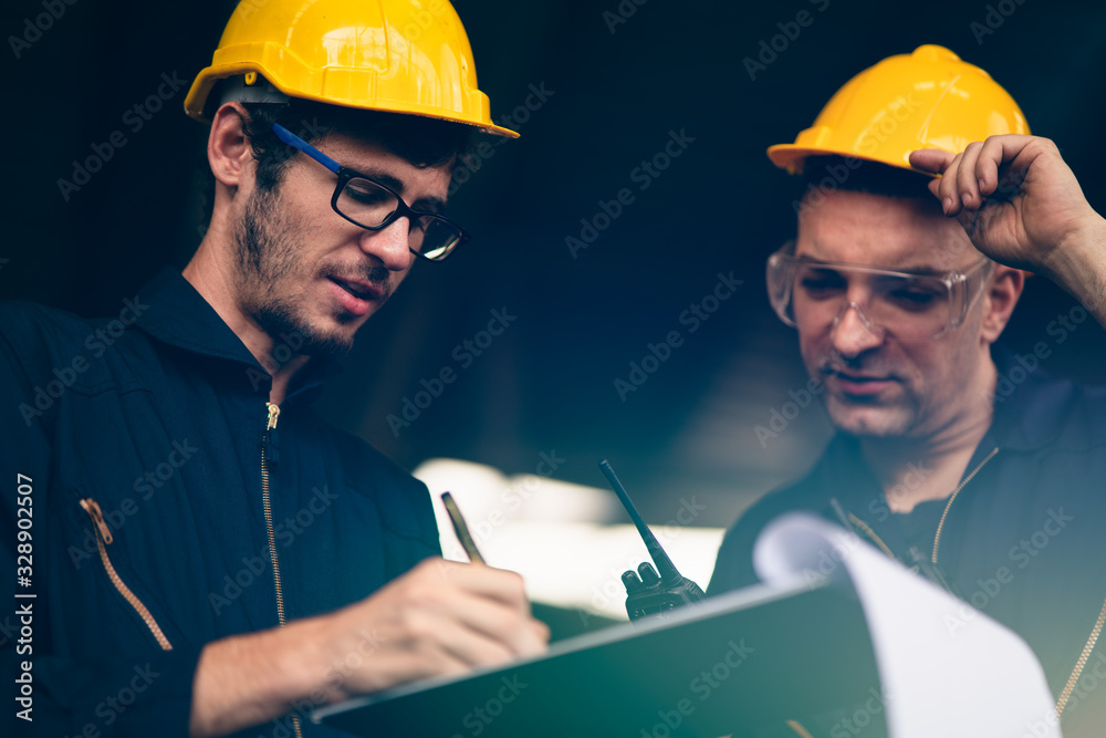 Scene of industrial workers wearing jump suit and yellow safety hat discussing the plan and operation while writing down the note on a clipboard, Concept manufacturer quality control procedure. Scene of industrial workers wearing jump suit and yellow safety hat discussing the plan and operation while writing down the note on a clipboard, Concept manufacturer quality control procedure.