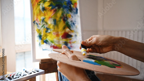 Cropped image of beautiful woman artist's hands while mixing color on the wooden color palette while sitting in front the painting canvas with modern art studio as background.