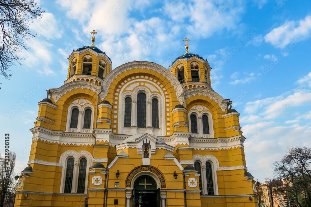 A view on the front facade of St Volodymyr's Cathedral. The cathedral is painted yellow, with blue and golden-domed rooftop. ON the side of the Cathedral there are trees. Overcast and cold day.