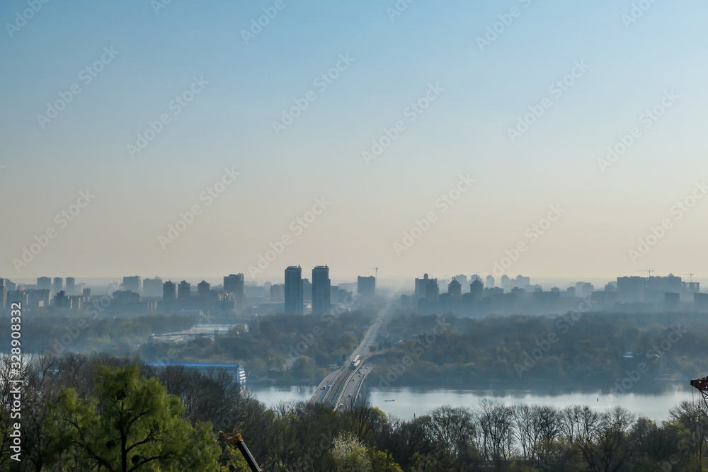 A panoramic city view on Kiev from a hill. In the middle there is a Dnieper river and a bridge joining two shores of the river. The city is shrouded in fog.