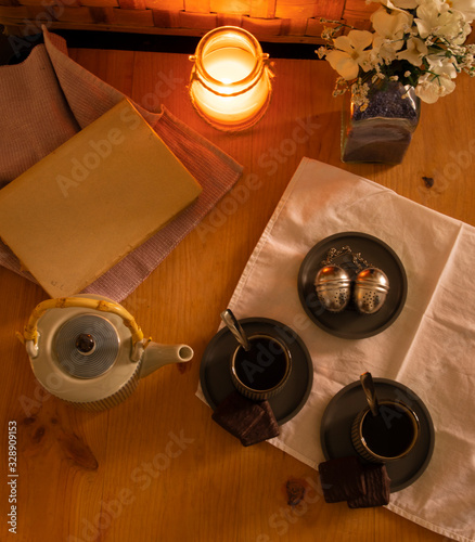 wooden table prepared for tea time. teapot, glasses of hot tea, book and flowers. in the light of the romantic candle giving a warm and welcoming tone to the scene. Chocolate cookies, and tea filter.