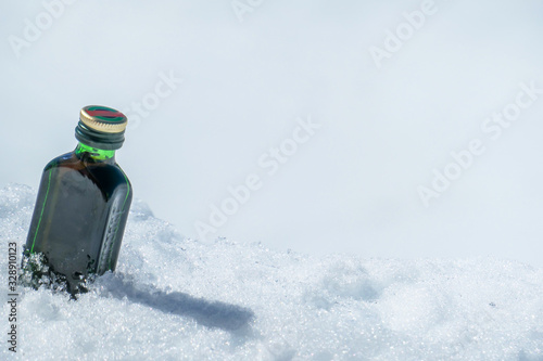 A little bottle of liquor placed on a fresh snow. The bottle has green color and a golden lid. The snow around the bottle is unspoiled. Apres ski in Alps.