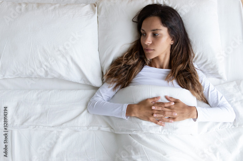 Top view of pensive young woman lying alone in cozy white bed thinking or pondering over relationships problem, depressed millennial girl relax in bedroom lost in thoughts after break up or divorce photo