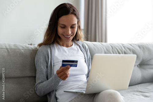 Smiling millennial girl sit on couch using laptop buying on Internet enter credit card details, happy young woman shopping online on computer make purchase on web, easy banking system service concept