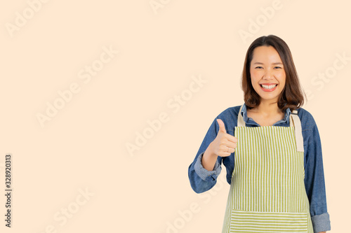close up young asian adult woman  (housewife)  wear apron  and raising hand thumb up for best score isolated on cream color background for good taste concept	