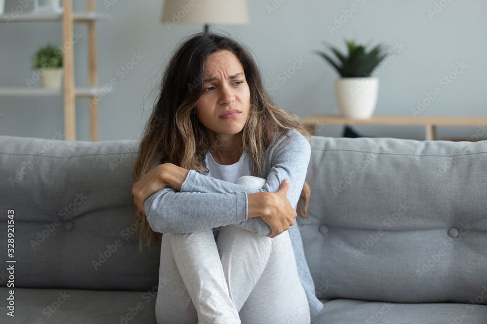 Sad millennial girl sit on couch feel lonely desperate look in distance morning yearning after breakup or divorce, unhappy young woman lost in thoughts suffer from depression or loneliness at home