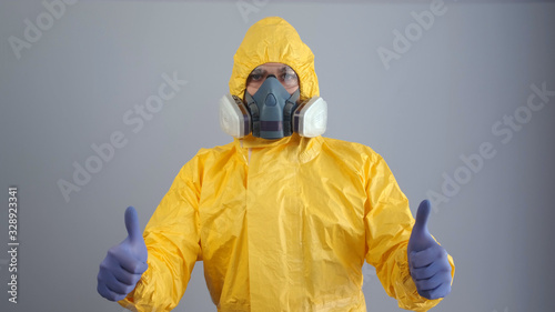 A man in yellow chemical protection suit and a respirator stands on a gray background and shows a thumbs up.