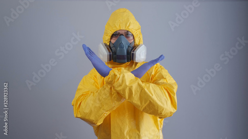 A man in yellow Hazmat suits and a respirator stands on a gray background and shows a stop sign with his hands. The man in crosses his arms over his chest, a sign to stop.