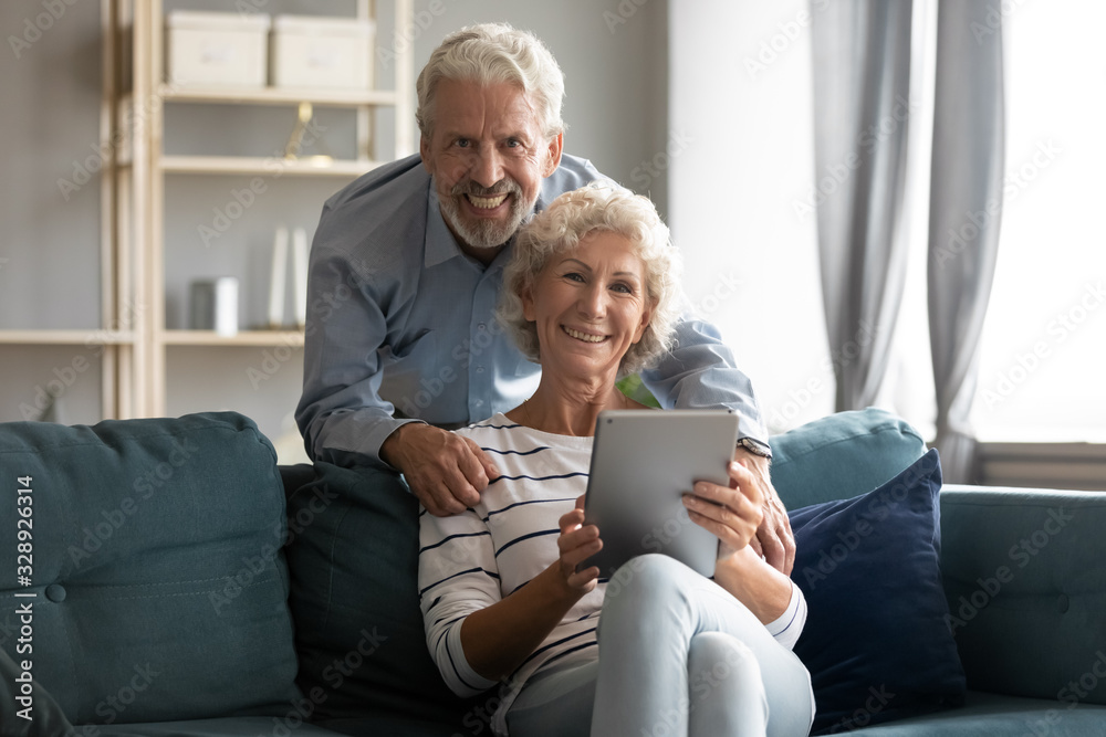 Portrait of smiling elderly 60s couple sit relax on cozy couch in living room using modern tablet together, happy old middle-aged 50s husband and wife look at camera browsing surfing internet on pad Portrait of smiling elderly 60s couple sit relax on cozy couch in living room using modern tablet together, happy old middle-aged 50s husband and wife look at camera browsing surfing internet on pad
