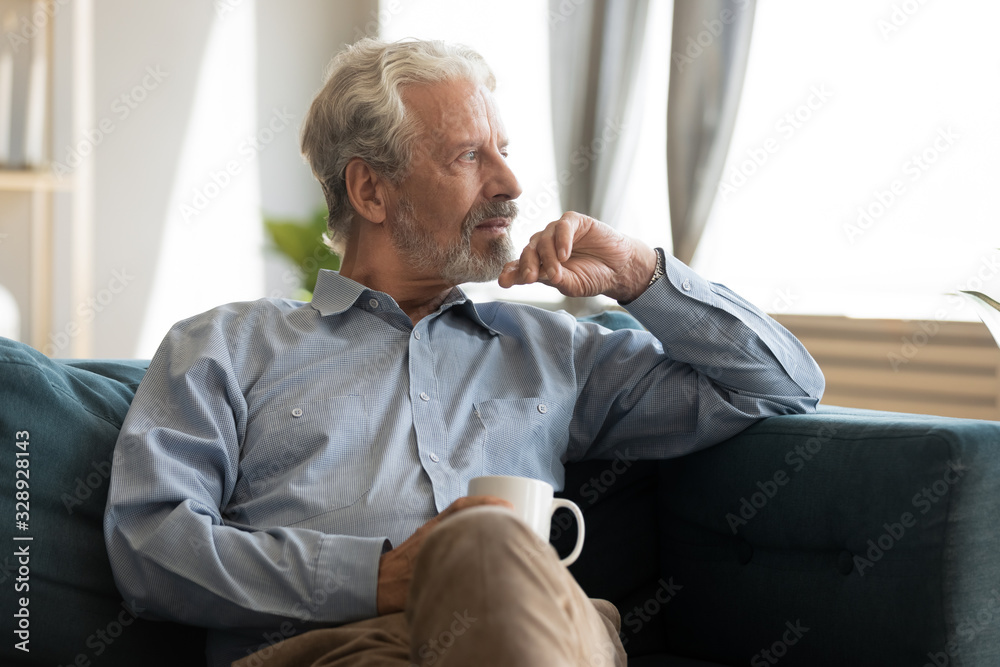 Pensive elderly 60s man sit relax on couch in living room drinking tea look  in window