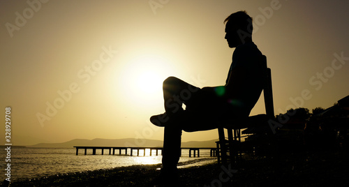Young sad depressed man sit alone on chair, looking at distant sea / seascape horizon. Time to go, say goodbye. Miss someone. Desire, hope to go far away. Unhappy boy feel lonely at seaside outdoor. 