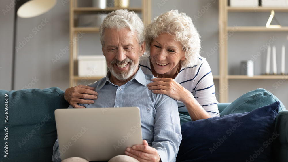 Happy elderly 50s husband and wife relax on comfy couch in living room watch funny video on laptop together, smiling old 60s couple rest on sofa at home have fun using modern computer gadget