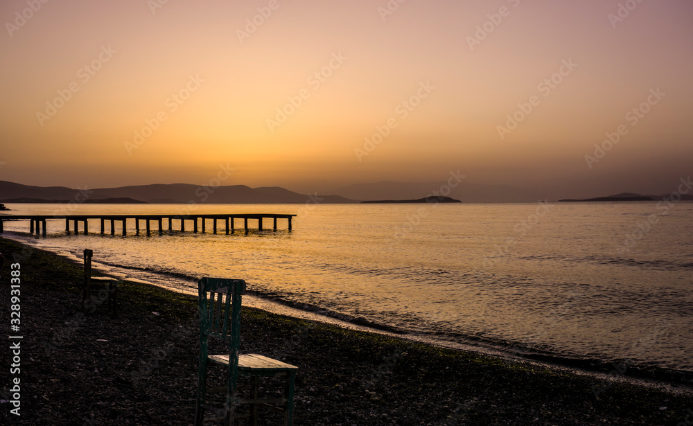 Empty wooden chairs on pebble beach during dramatic sunset. Summer is coming and outdoor / outside conceptual photo. Amazing sunset over mountains & ocean horizon at weekend. Sunny day at outdoor cafe