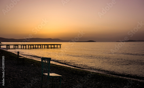 Empty wooden chairs on pebble beach during dramatic sunset. Summer is coming and outdoor   outside conceptual photo. Amazing sunset over mountains   ocean horizon at weekend. Sunny day at outdoor cafe