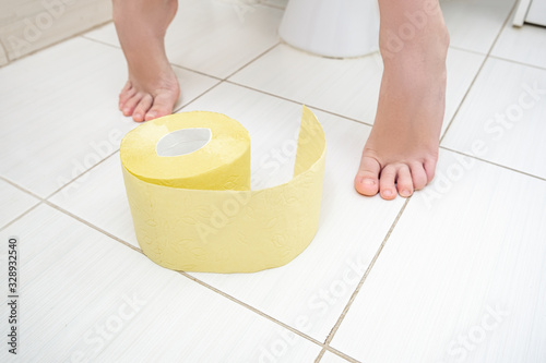 Cropped legs of a boy sitting on a toilet with a roll of yellow toilet paper on a floor, digestive problems and difficulties in toilet concept