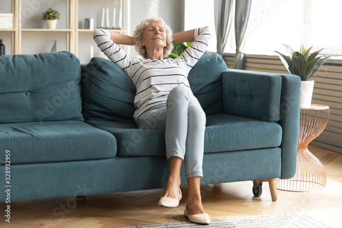Calm elderly 60s woman rest on cozy sofa in living room hands overhead eyes closed, peaceful mature 50s wife relax on comfortable couch at home take nap daydream, stress free, peace concept