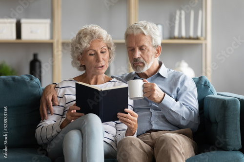 Happy middle-aged 50s husband and wife sit relax on couch in living room reading book drinking tea together, calm elderly 60s couple booklovers rest at home enjoy story novel on weekend