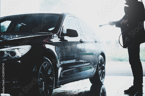 Fototapeta Naklejka Na Ścianę i Meble -  Silhouette of a man washing his black car outdoors in a carwash station, using water jet and high pressured water stream
