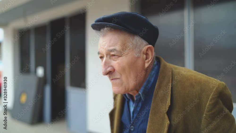 Portrait of senior man pensioner looking around while standing alone outdoor wearing jacket and hat cap in front of house Portrait of senior man pensioner looking around while standing alone outdoor wearing jacket and hat cap in front of house