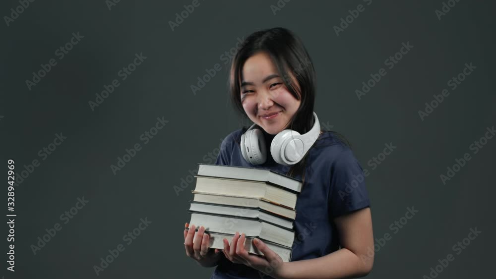 Asian student girl with headphones on grey background in studio holds stack of university books from library. Woman smiles, she is happy to graduate. Asian student girl with headphones on grey background in studio holds stack of university books from library. Woman smiles, she is happy to graduate.