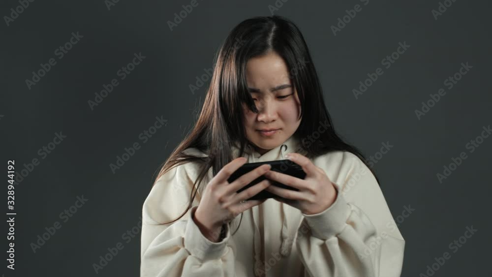 Girl with asian appearance playing game on smartphone on grey studio wall. Using modern technology - apps, social networks Girl with asian appearance playing game on smartphone on grey studio wall. Using modern technology - apps, social networks